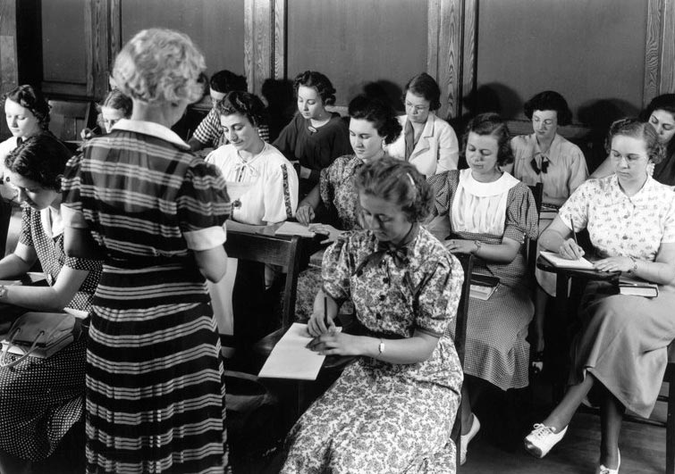 Women in the 40s/50s wear take notes in a class led by an elderly woman. The picture is black and white.