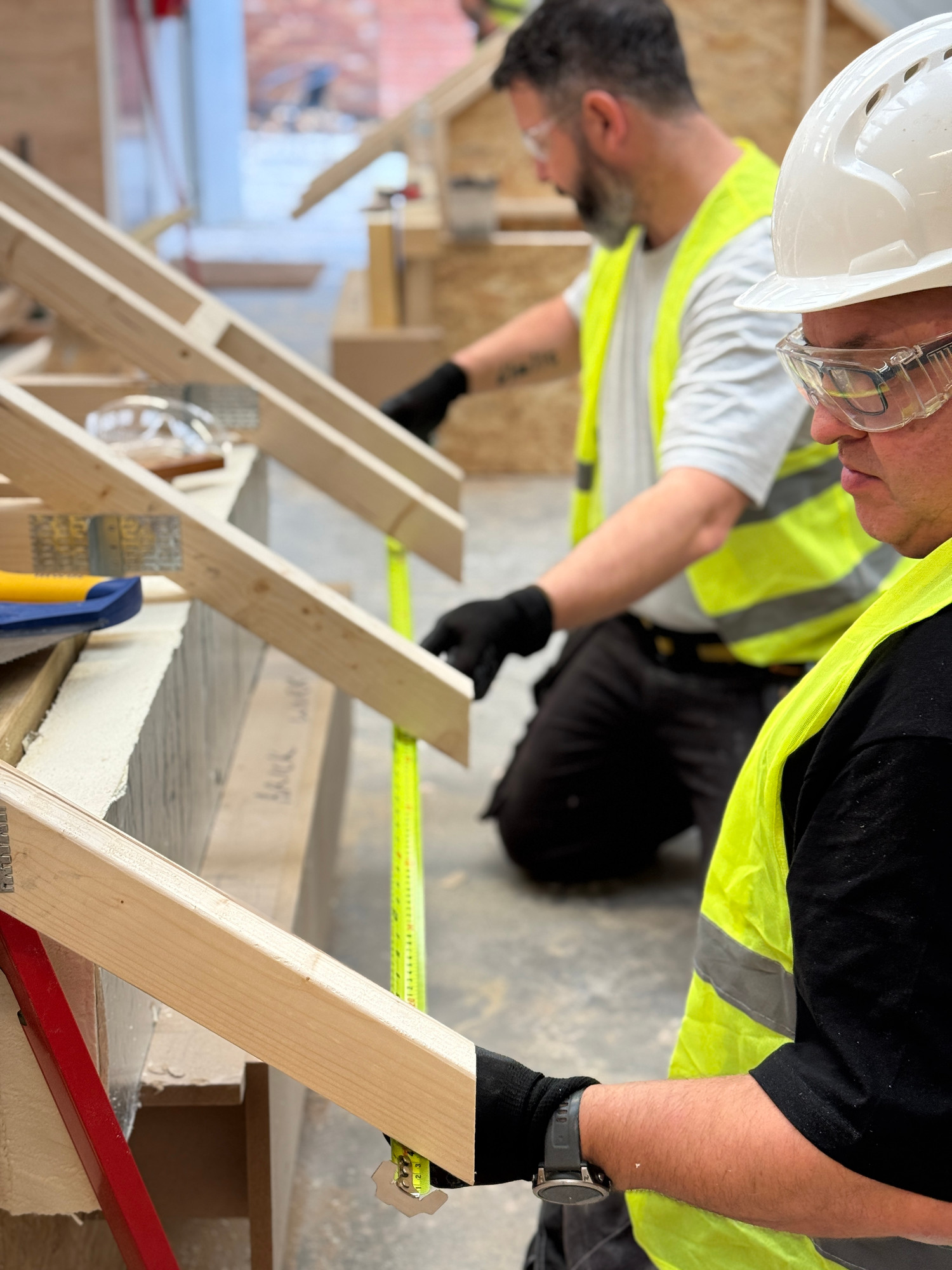 Two people measure wood with a tape measure. They wear hard hats and protective glasses and fluorescent jackets