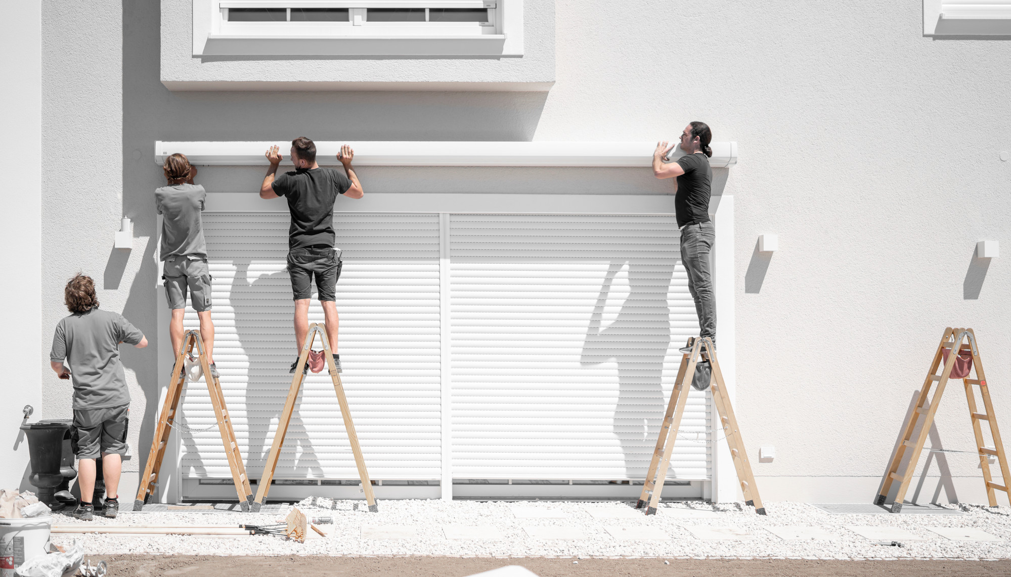 3 men work across three ladders putting up an awning above a garage door. One man instructs from the ground.
