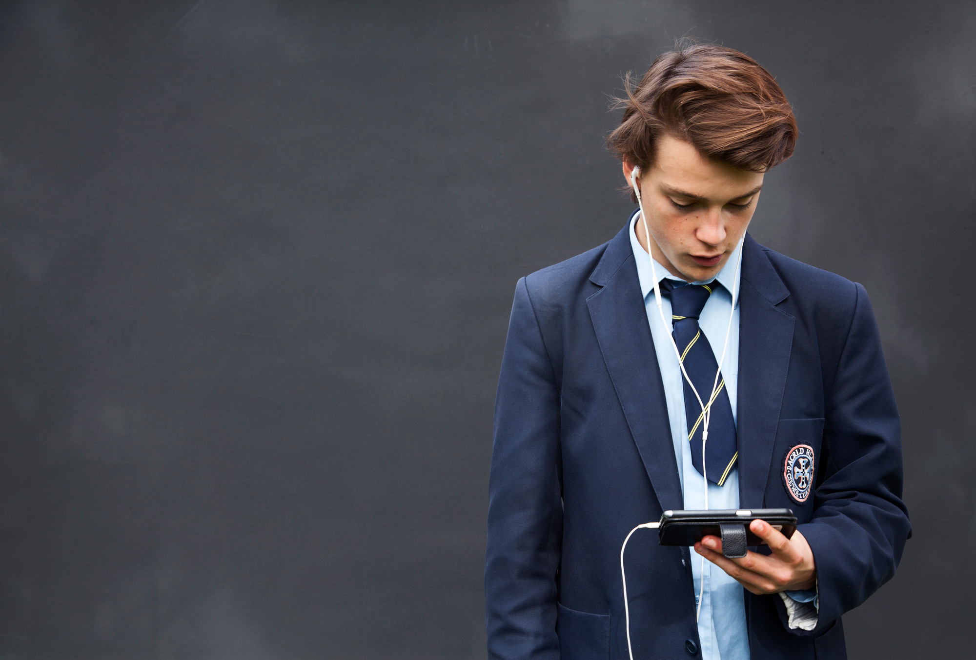 A boy in school uniform watches his smartphone whilst wearing white in earphones with a white wire.