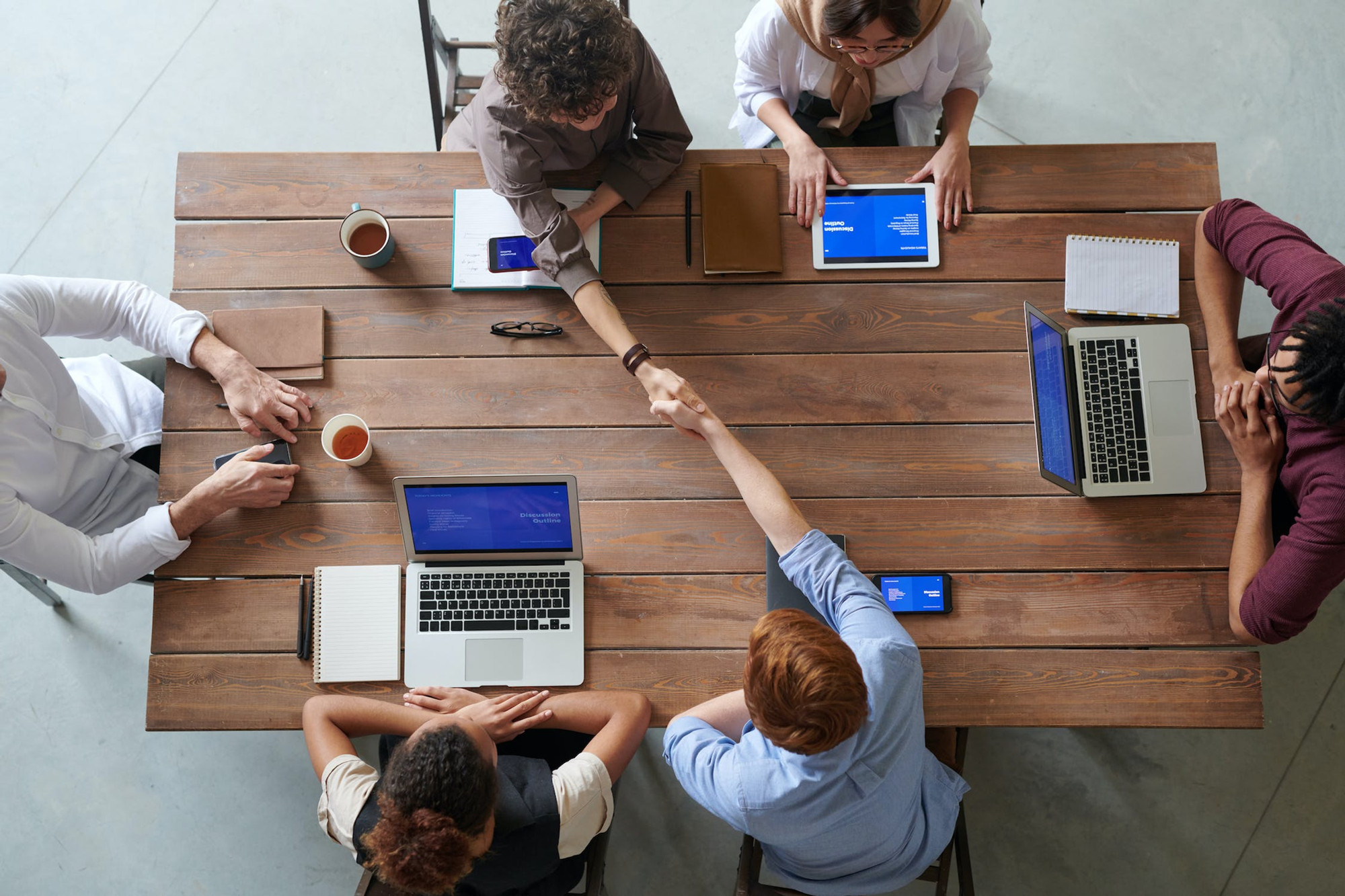 A Birds Eye View of a co working table where a group of people work on their laptops. Two people shake hands across the table