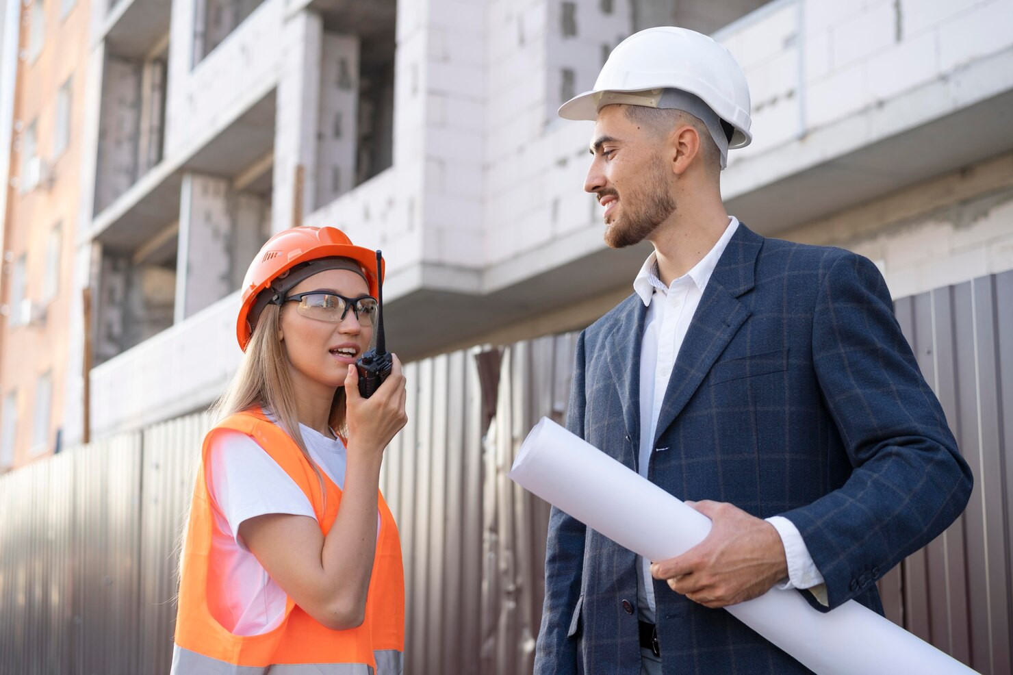 A woman in a hard hat and fluorescent jacket and white t shirt speaks into a walkie talkie. Next to her, a man in a suit, also wearing a hard hat holding a large, rolled up piece of paper. They stand in front of a construction site.