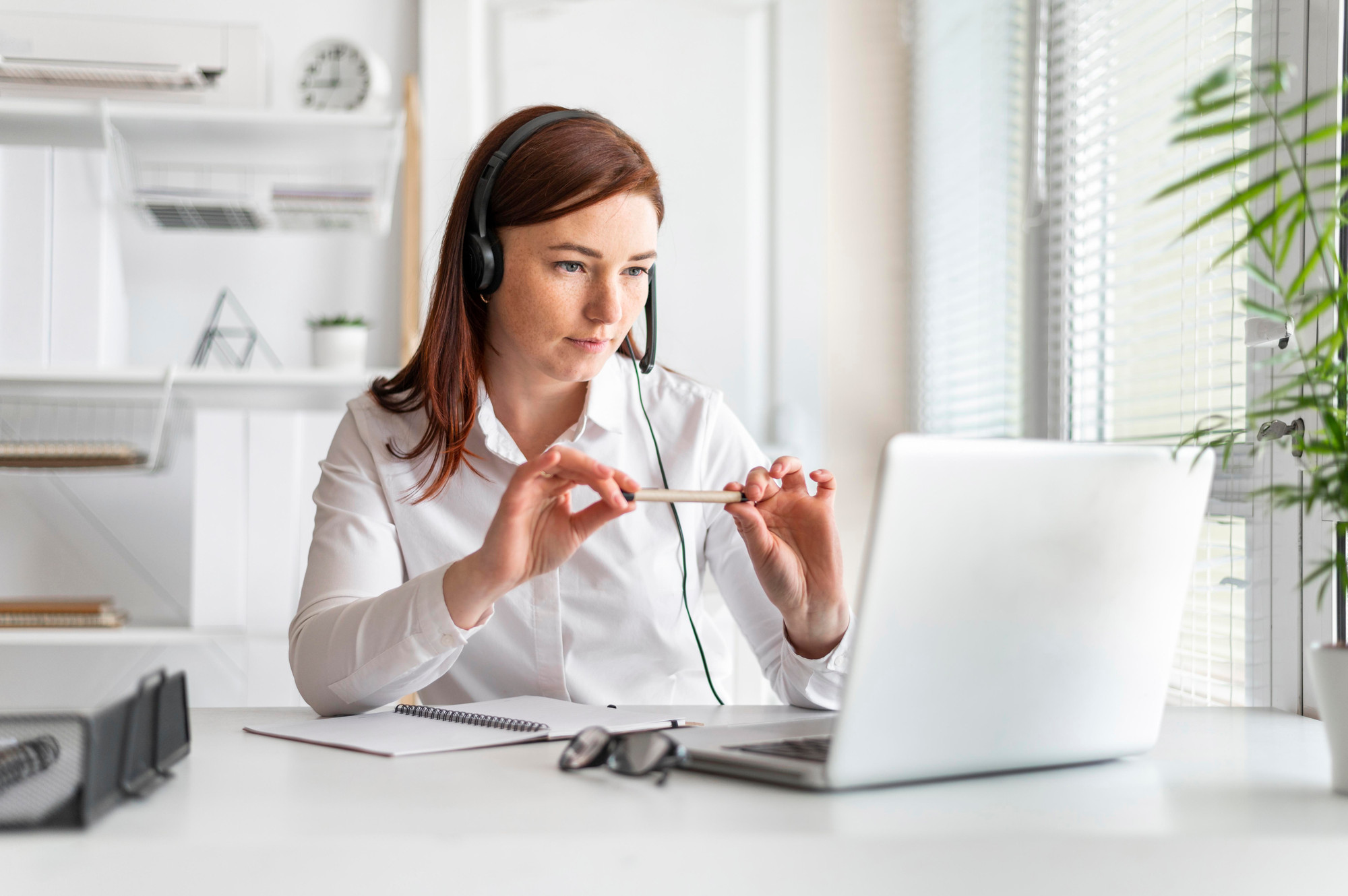 A woman wearing a headset and holding a pen sits attentively looking at her laptop in a pristine white office next to a green plant. An empty notebook is open in front of her.