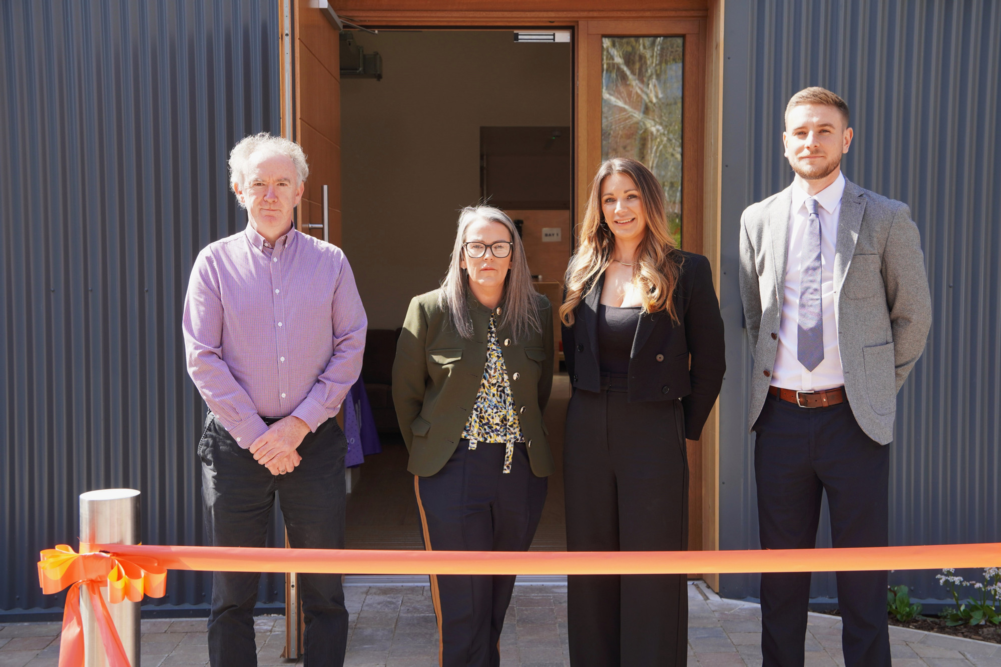 4 of our team stand in front of the building waiting to cut an orange ribbon and declare it open.