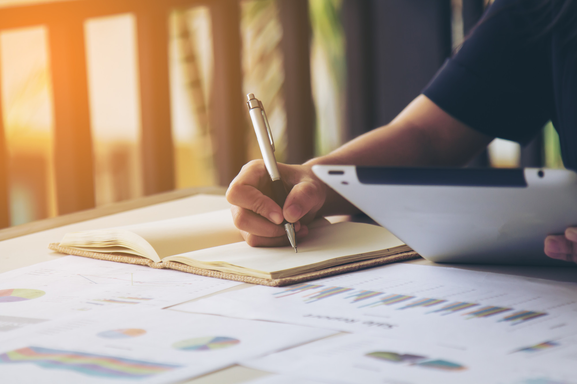 A hand takes notes with a pen and a notebook whilst holding a tablet device.