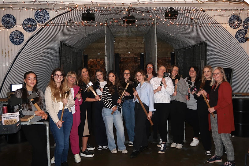 A team of women in smart casual wear pose holding axes at an axe throwing centre. They're having a team social.
