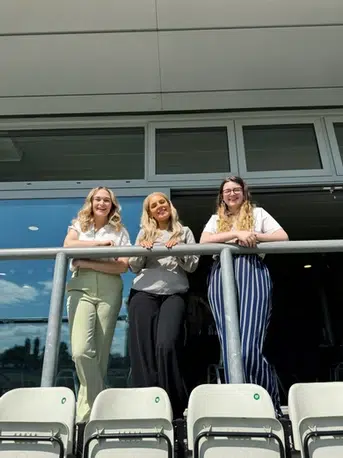 3 women pose from the standing area looking down over an outdoor stadium's seats. They all smile.
