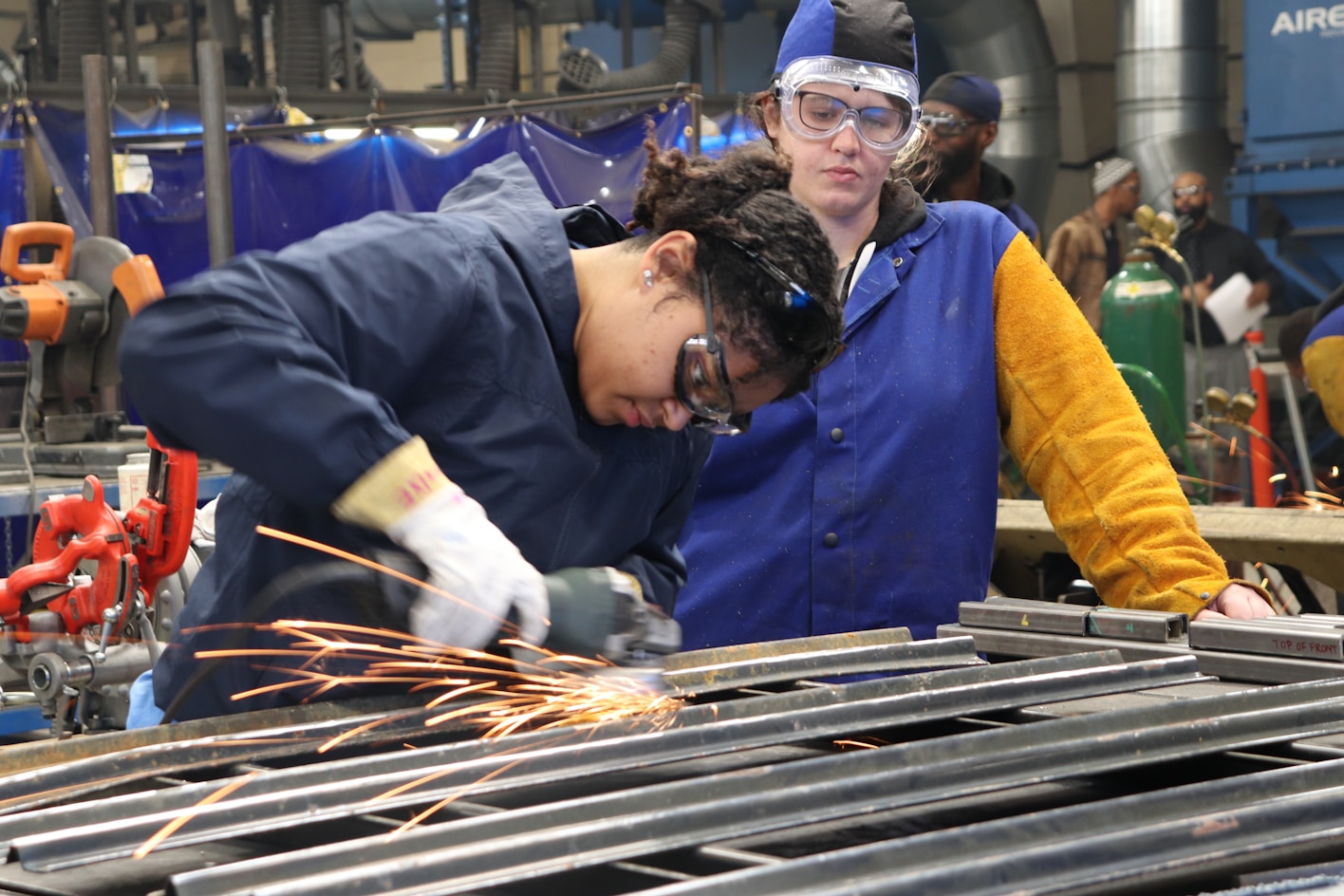 Two female students in protective clothes and goggles perform metal work. Sparks fly from the metal grinder meeting the worked on metal.