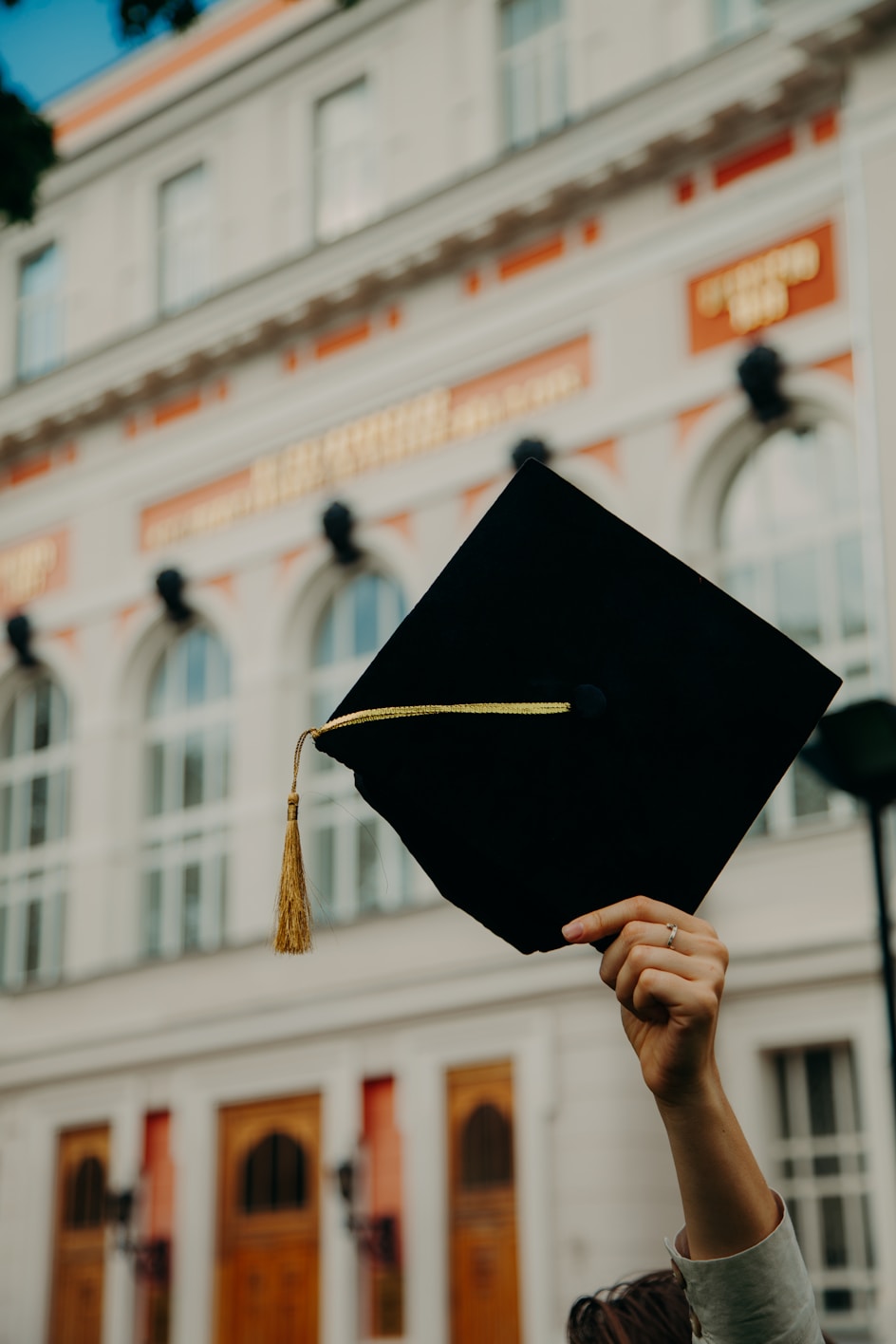 A close up of a black graduation hat being held high in front of a grand looking building.