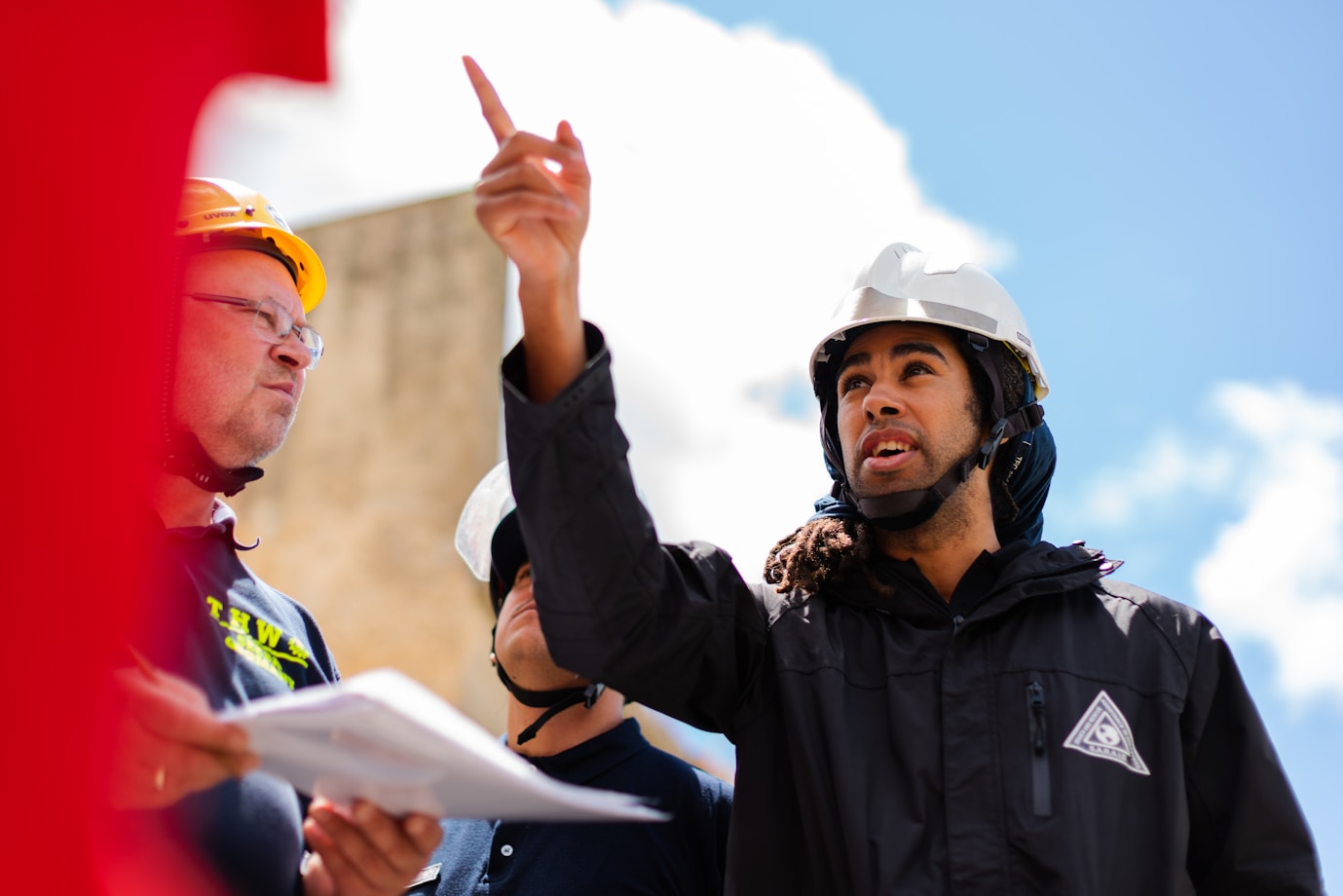 A student in a hard hat points upward. A tutor listens to him whilst holding a piece of paper and also wearing a hard hat. They are outside.
