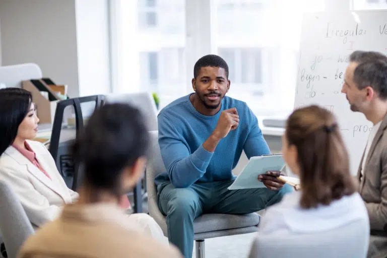 A man refers to a clipboard in his hands whilst talking to 4 other people in a circle. Behind him is a white board that indicates 'Irregular Verbs'
