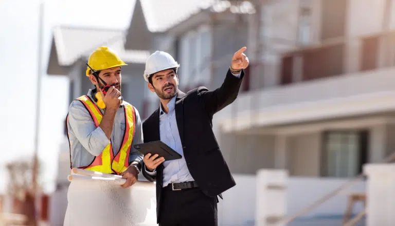 A manager of a construction site points out in a collaborative manor with his site working colleague who speaks into a walkie talkie.