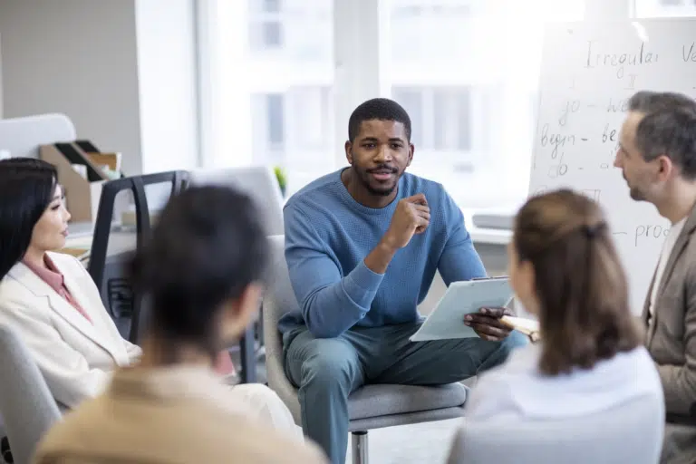 A man refers to a clipboard in his hands whilst talking to 4 other people in a circle. Behind him is a white board that indicates 'Irregular Verbs'