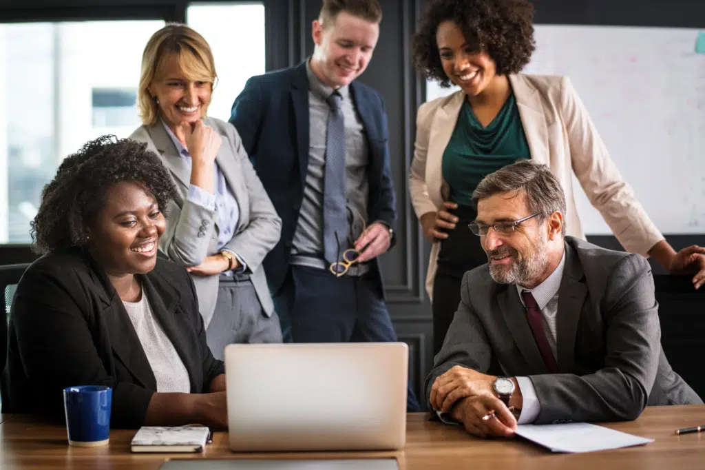 A group of workers look happy and pleased around a laptop in an office. They all wear formal office wear.
