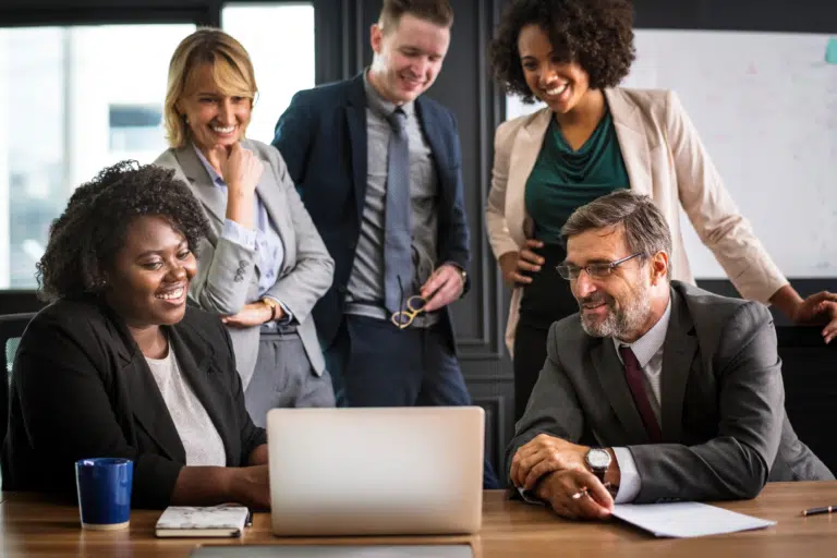 A group of workers look happy and pleased around a laptop in an office. They all wear formal office wear.