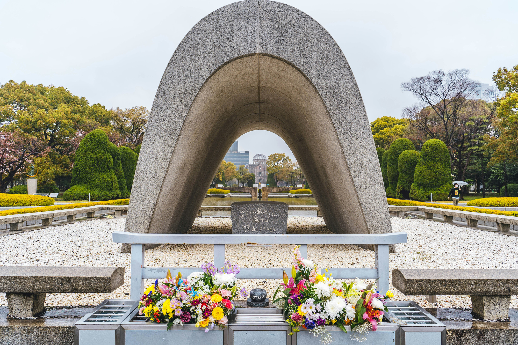 A curved, concrete monument structure, framed by concrete benches and gravel. This is clearly in a public park surrounded by greenery on a cloudy day.