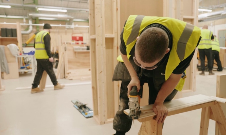 two learners in a construction workshop setting, one is cutting wood up front and the other is walking in the background, both wearing PPE